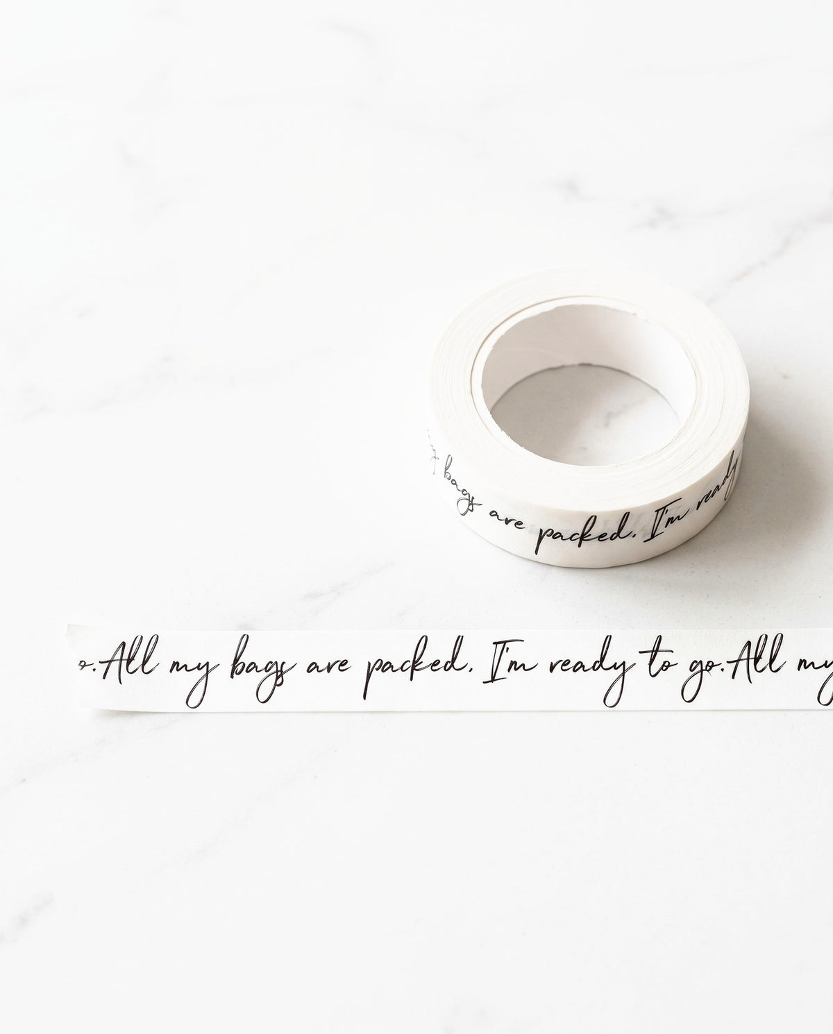 Roll of All My Bags Are Packed washi tape with a strip pulled out, on white marble