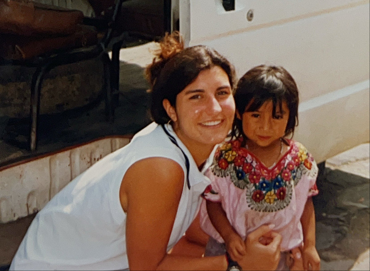 Sairey, Founder of Trouvaille Collective,  in a white sleeveless top crouches beside a young child in Guatemala in front of a white vehicle door, with a worn brown bench visible inside the vehicle.