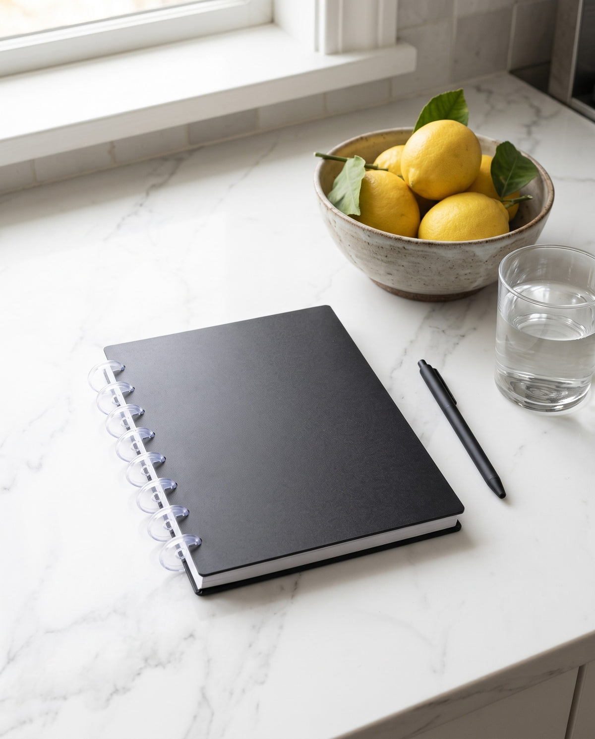 Black notebook cover set on a half letter discbound planner with 8 clear discs, on a white marble kitchen counter with a bowl of lemons