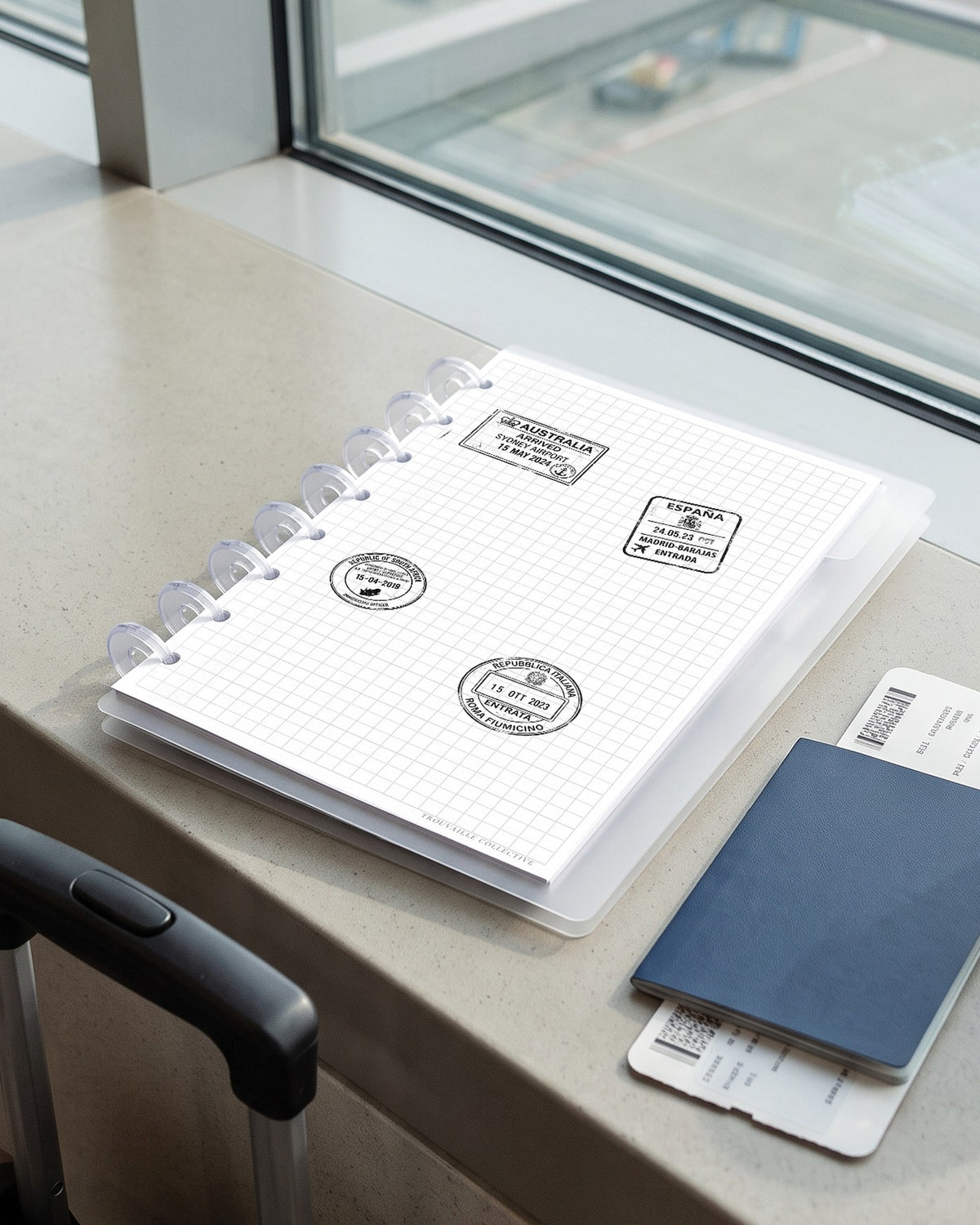 Passport Stamps clear stickers applied to a planner page in a half letter discbound planner on an airport gate window ledge