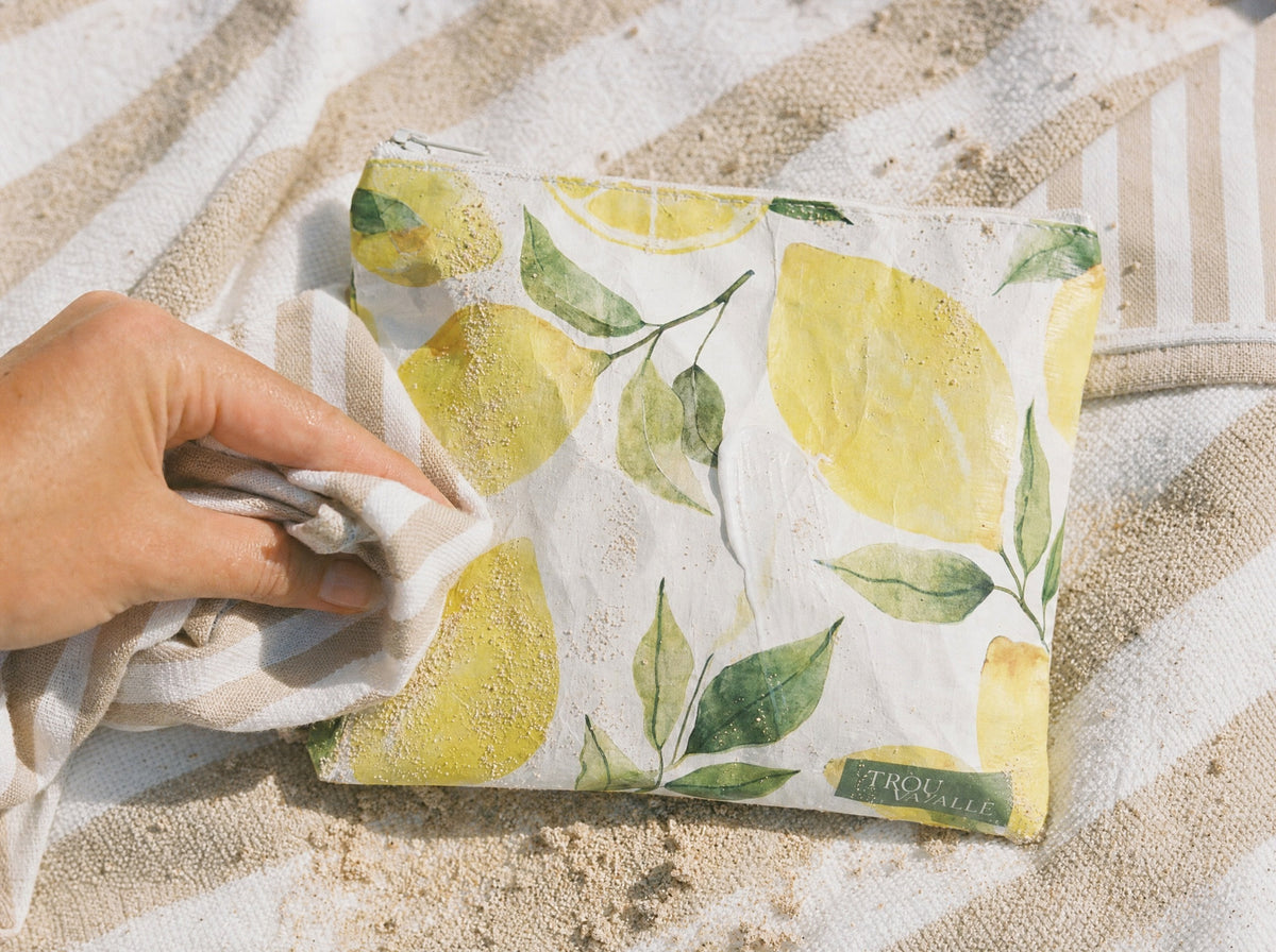 A sandy Trouvaille water-resistant accessory bag being wiped clean with the corner of a beach towel